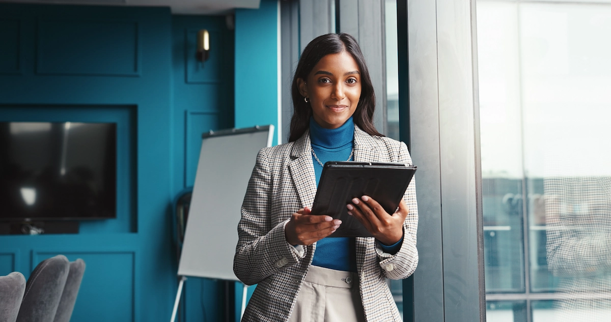 Confident professional woman holding a tablet, standing near a window in a bright, modern office.